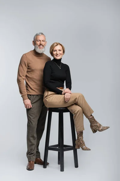 Sonriente hombre con estilo mirando a la cámara cerca de la esposa sentada en la silla sobre fondo gris - foto de stock