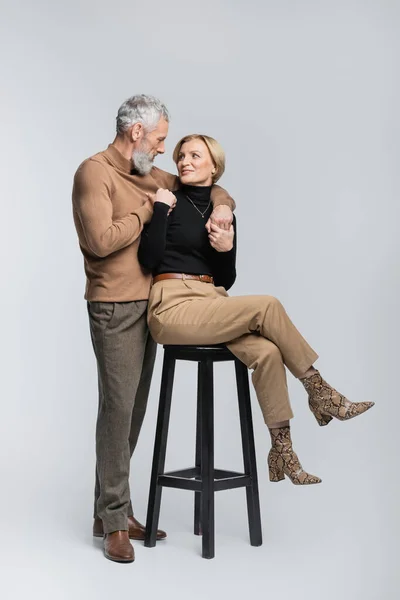 Mature man hugging stylish wife sitting on chair on grey background — Stock Photo