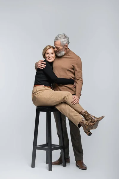 Alegre elegante mujer abrazando maduro marido mientras sentado en silla en gris fondo - foto de stock