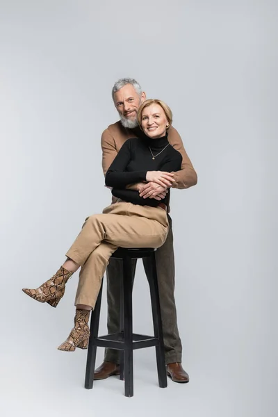 Позитивный зрелый мужчина, обнимающий модную жену, сидящую на стуле на сером фоне — стоковое фото