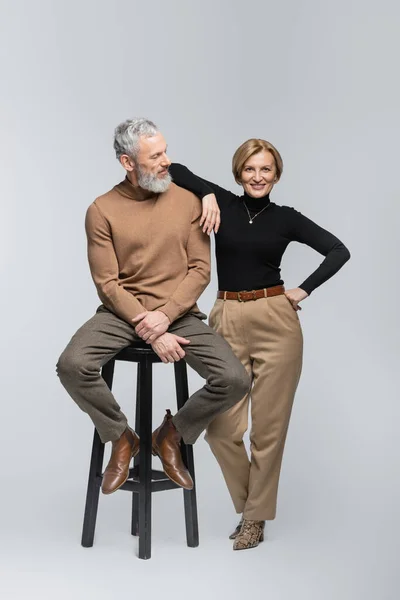 Sonriente mujer madura posando cerca de marido elegante sentado en la silla sobre fondo gris - foto de stock