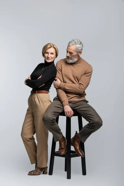 Full length of stylish woman crossing arms near mature husband sitting on chair on grey background — Stock Photo