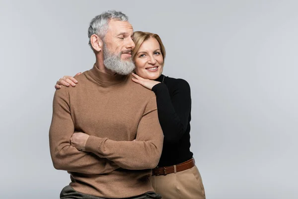 Smiling mature woman in turtleneck hugging husband and looking at camera isolated on grey — Stock Photo