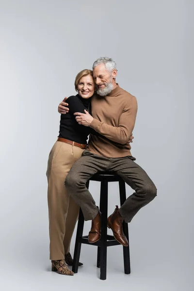 Sonriente mujer madura abrazando marido sentado en la silla sobre fondo gris - foto de stock