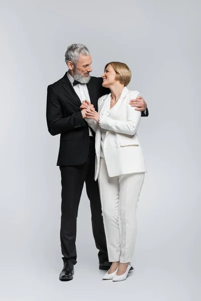 Full length of grey haired groom hugging bride in suit on grey background — Stock Photo