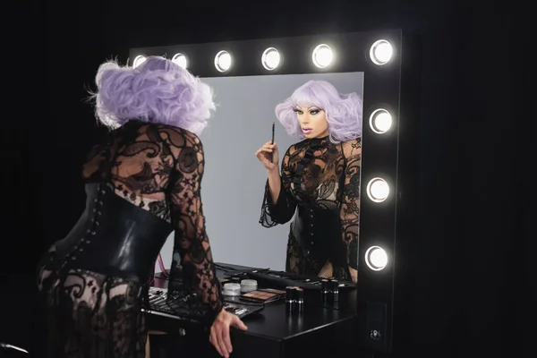 Drag queen em peruca violeta e vestido de renda preta segurando escova cosmética perto do espelho — Fotografia de Stock