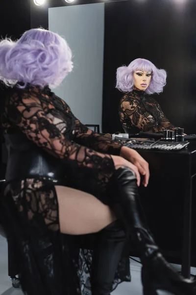 Drag queen glamour en perruque violette et robe en dentelle noire assise avec jambes croisées près du miroir — Photo de stock