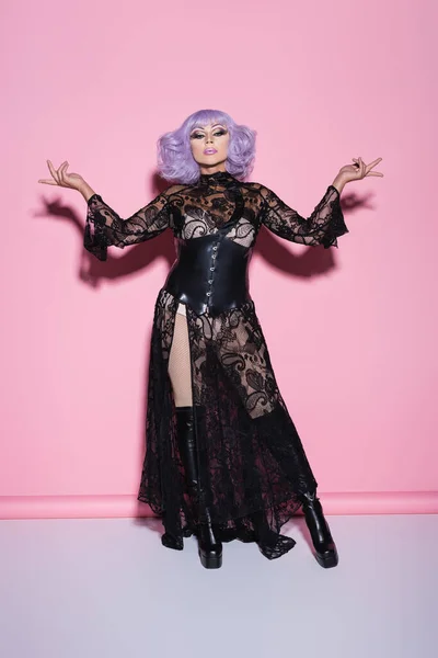 Transgender person in black lace dress and violet wig standing with open arms on pink — Stock Photo