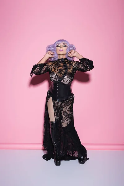 Visão de comprimento total do excêntrico drag queen em vestido de renda preta olhando para a câmera enquanto em pé sobre rosa — Fotografia de Stock