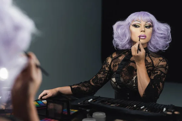 Drag queen en perruque violette et robe en dentelle noire appliquant le maquillage près du miroir — Photo de stock