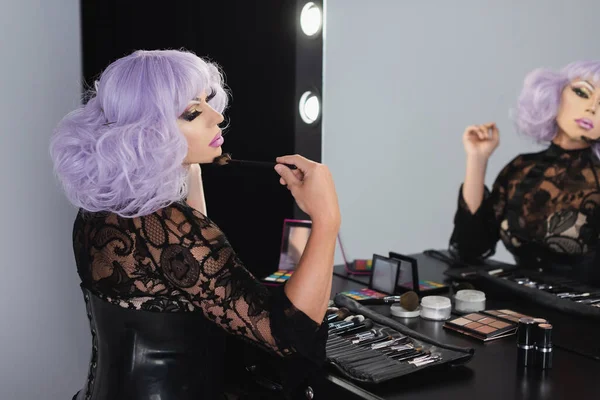 Drag queen em peruca violeta aplicando pó facial com escova cosmética perto do espelho — Fotografia de Stock