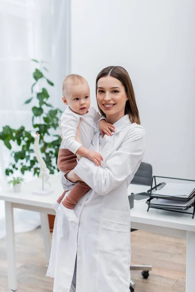 Positive family doctor in white coat holding baby boy and looking at camera in clinic — Stock Photo