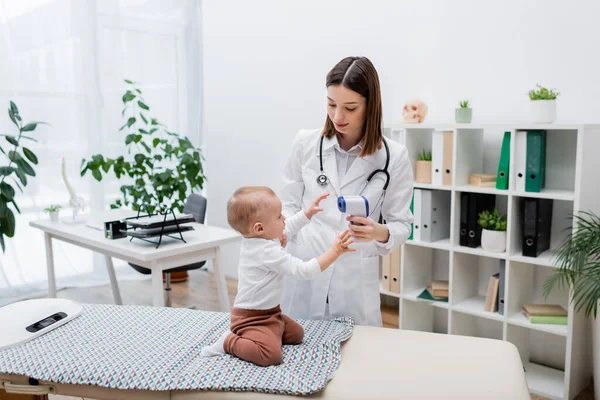 Junger Hausarzt hält berührungsloses Pyrometer nahe Baby auf Medizinercouch in Klinik — Stockfoto