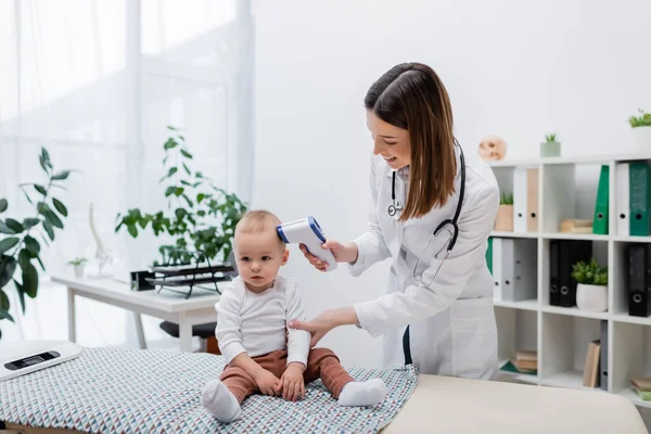 Positive family doctor checking temperature of baby boy with pyrometer in clinic — Stock Photo