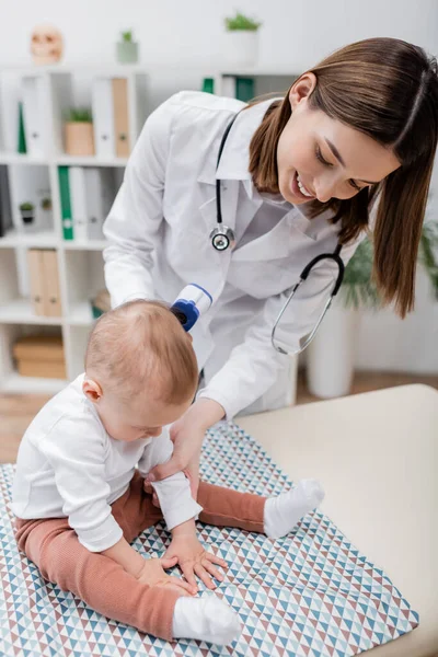 Joyeux médecin de famille tenant pyromètre près du bébé sur le canapé médical à la clinique — Photo de stock