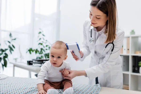 Baby boy looking at camera near family doctor with pyrometer in clinic — Stock Photo