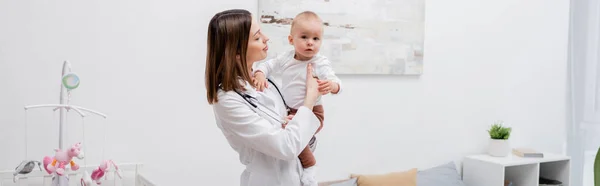 Young family doctor in white cot holding baby boy at home, banner — Stock Photo