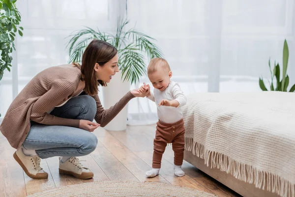 Happy mother holding hand of smiling baby near bed at home — Stock Photo