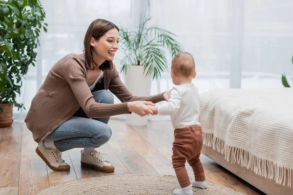 Positive young woman holding hands of baby boy in bedroom — Stock Photo