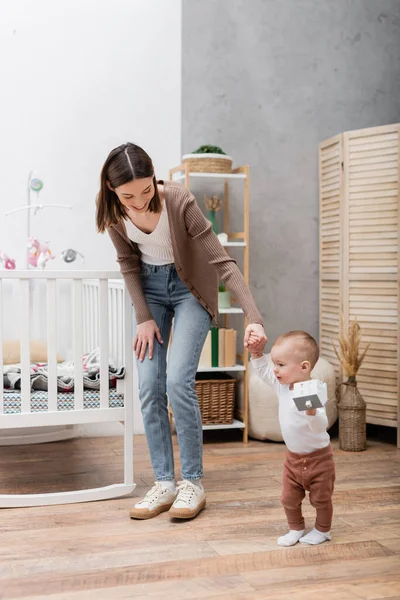Smiling woman holding hand of baby boy with toy house in bedroom — Stock Photo
