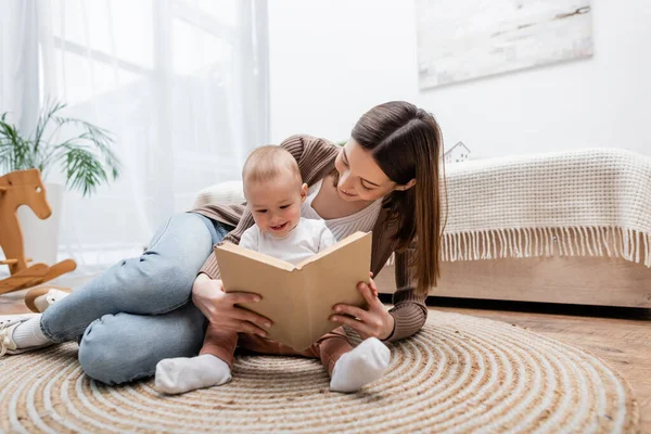 Jeune maman livre de lecture avec bébé fils sur le sol dans la chambre — Photo de stock