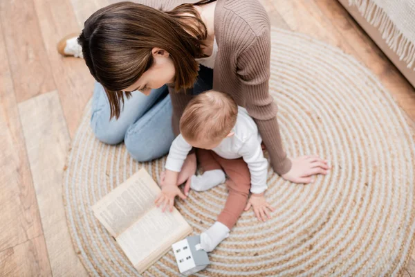 Overhead view of woman reading book near blurred baby son on floor at home — Stock Photo