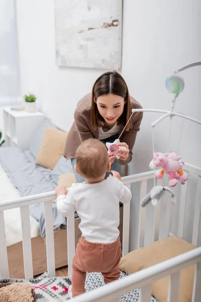 Morena mãe segurando brinquedo perto do filho no berço no quarto — Fotografia de Stock