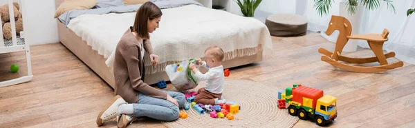 Woman looking at baby son holding bag with building blocks in bedroom, banner — Stock Photo