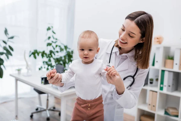 Family doctor in white coat holding hands of infant patient in clinic — Stock Photo