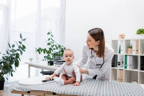 Smiling pediatrician holding baby near scales on medical couch in hospital — Stock Photo