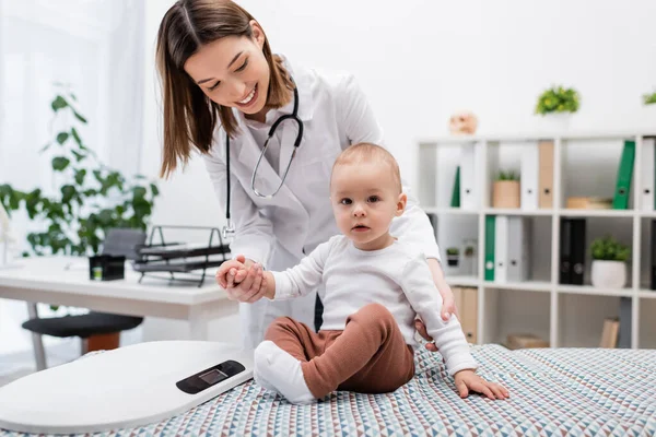 Happy family doctor looking at child near scales on medical couch in clinic — Stock Photo