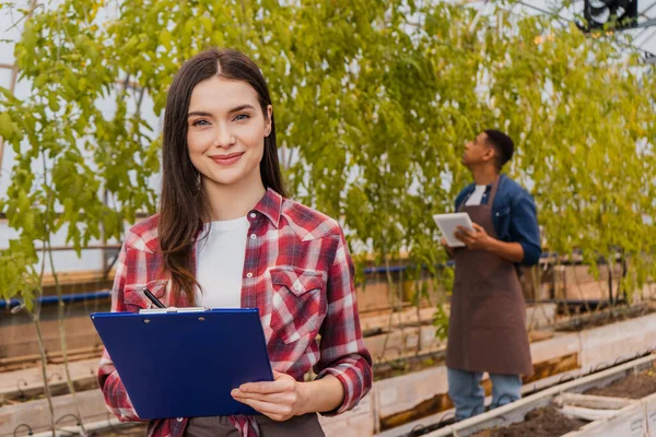 Smiling farmer looking at camera while holding clipboard near blurred african american colleague in greenhouse — Stock Photo