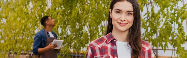 Jungbauer lächelt in die Kamera neben verschwommenem afrikanisch-amerikanischem Kollegen mit Klemmbrett im Gewächshaus, Banner — Stockfoto