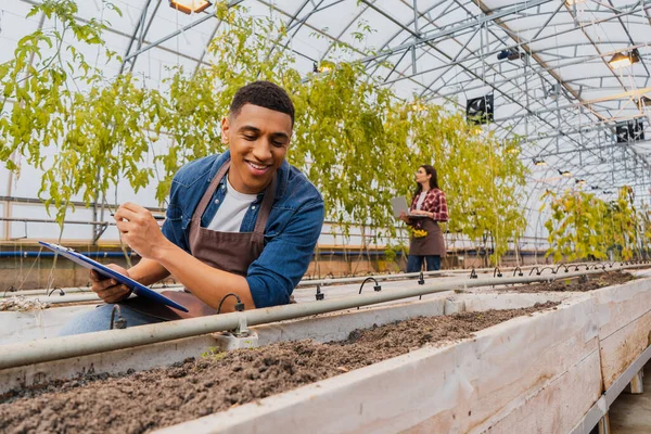 Happy african american farmer holding clipboard near ground in greenhouse — Stock Photo