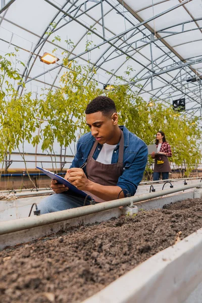 African american farmer writing on clipboard near ground in greenhouse — Stock Photo