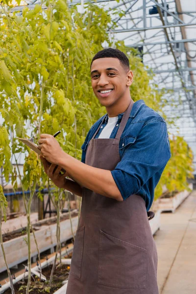 Positive african american farmer in apron writing on clipboard near plants in greenhouse — Stock Photo
