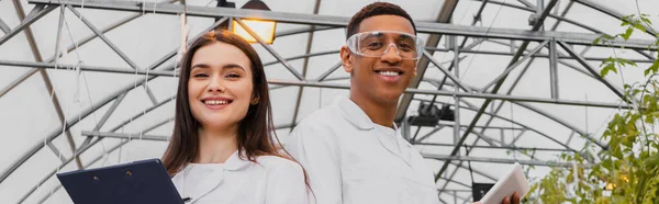 Low angle view of cheerful interracial botanists with clipboard and digital tablet looking at camera in greenhouse, banner — Stock Photo