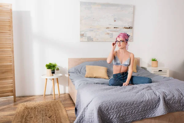 Молодая женщина с розовыми волосами регулируя очки и сидя на кровати — стоковое фото