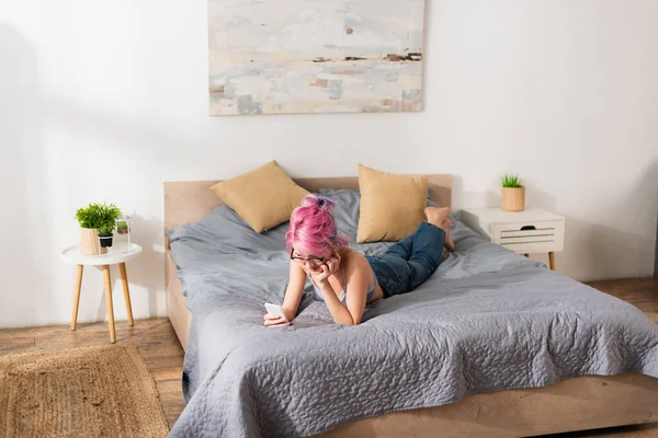 Young woman with pink hair in bra and jeans using smartphone while lying on bed — Stock Photo