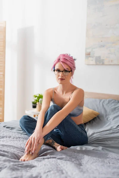 Tattooed young woman with pink hair sitting in bra and jeans on bed — Stock Photo