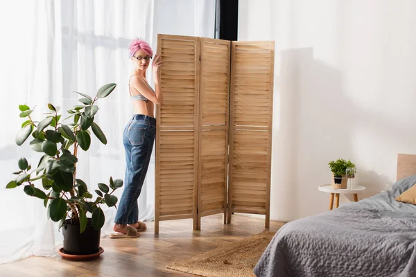 Tutta la lunghezza della giovane donna con i capelli tinti in piedi in reggiseno e jeans vicino schermo pieghevole — Foto stock