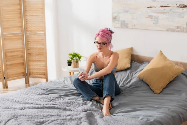 Junge Frau mit gefärbten Haaren sitzt in Jeans und BH während sie Nachrichten auf dem Smartphone sendet — Stockfoto