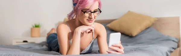 Fröhliche junge Frau mit gefärbten Haaren, die auf dem Bett liegt, während sie auf dem Smartphone Nachrichten sendet, Banner — Stockfoto