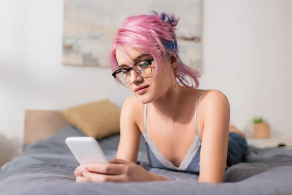 Jovem bonita com cabelo tingido deitado na cama enquanto conversa no smartphone — Fotografia de Stock