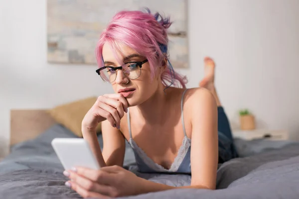 Pensive young woman with dyed hair lying on bed while using smartphone — Stock Photo