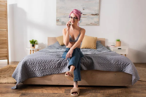 Happy young woman with dyed hair sitting in jeans and bra on bed while talking on smartphone — Stock Photo