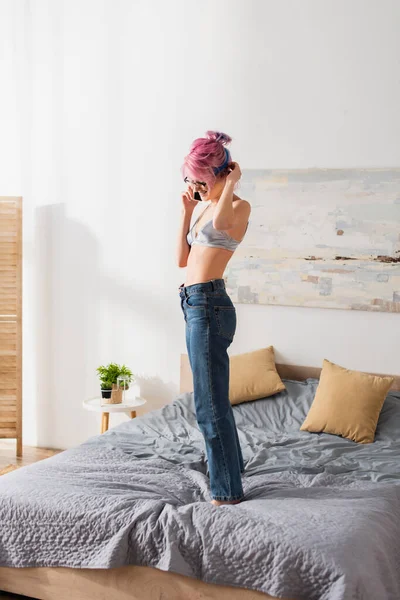 Улыбающаяся молодая женщина с окрашенными волосами, стоящая на кровати, разговаривая по смартфону — стоковое фото