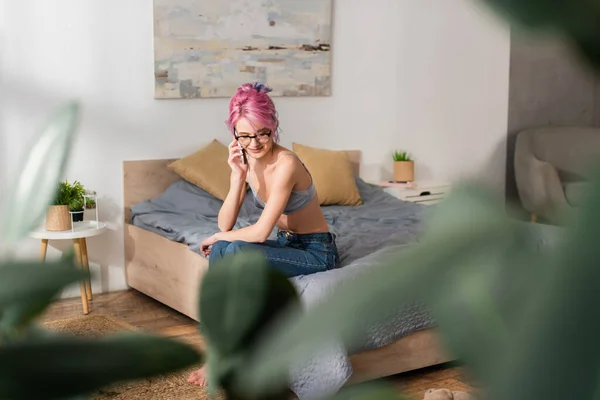 Lächelnde junge Frau mit gefärbten Haaren, die auf dem Bett sitzt, während sie mit dem Handy spricht — Stockfoto