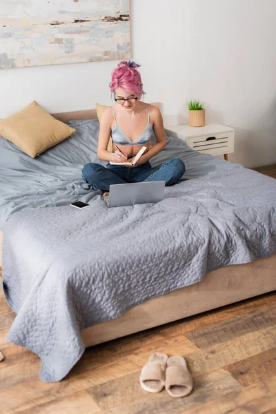 Mujer joven con el pelo teñido haciendo notas en el cuaderno mientras estudia en línea en casa - foto de stock