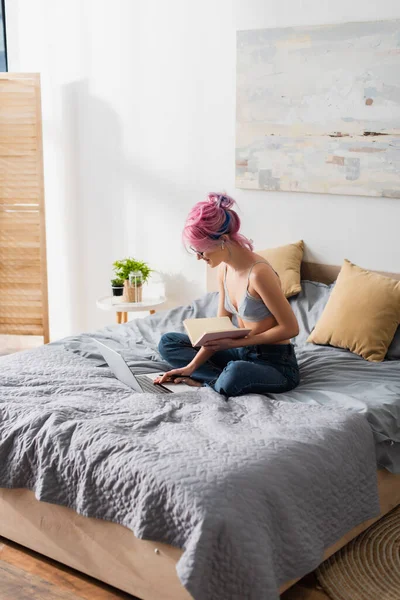 Femme gaie avec les cheveux teints tenant carnet tout en utilisant un ordinateur portable et étudier en ligne à la maison — Photo de stock